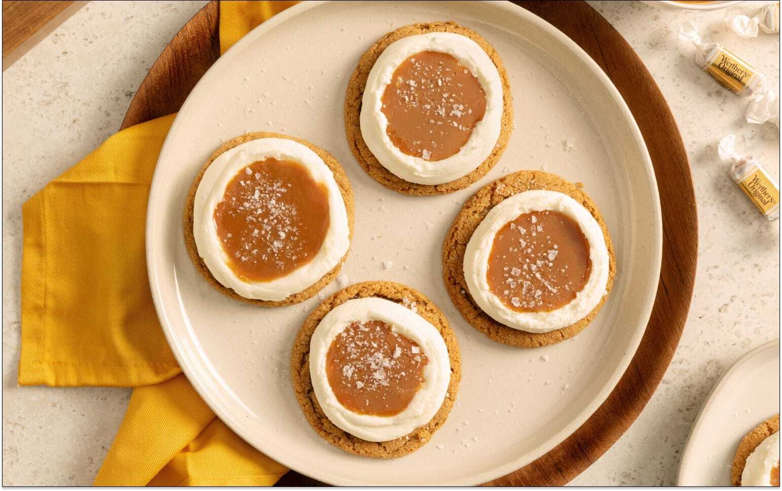 Four round biscuits topped with a ring of white icing and a caramel centre, lightly sprinkled with sugar, arranged on a cream plate with a yellow napkin underneath.