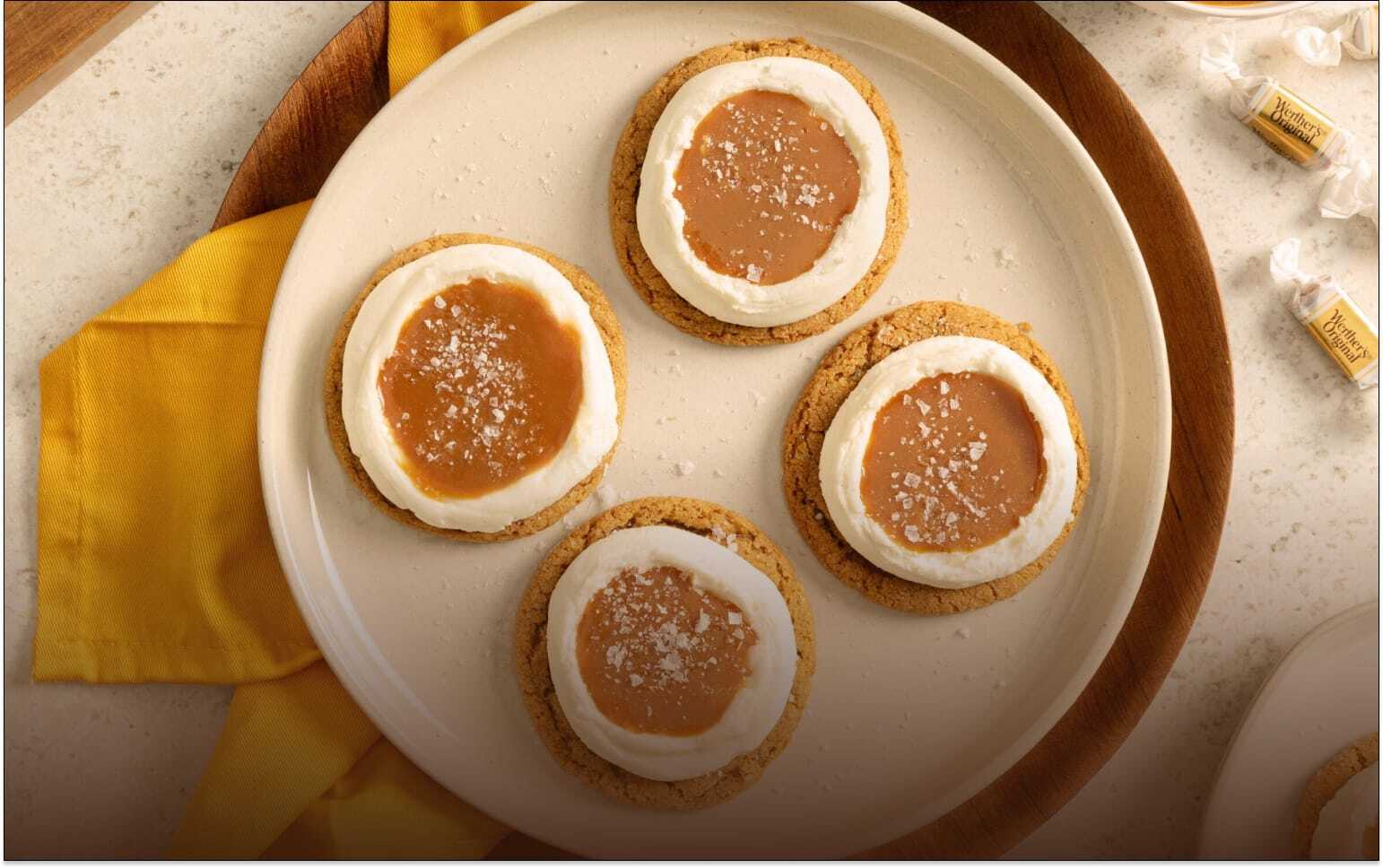 Four round biscuits topped with a ring of white icing and a caramel centre, lightly sprinkled with sugar, arranged on a cream plate with a yellow napkin underneath.