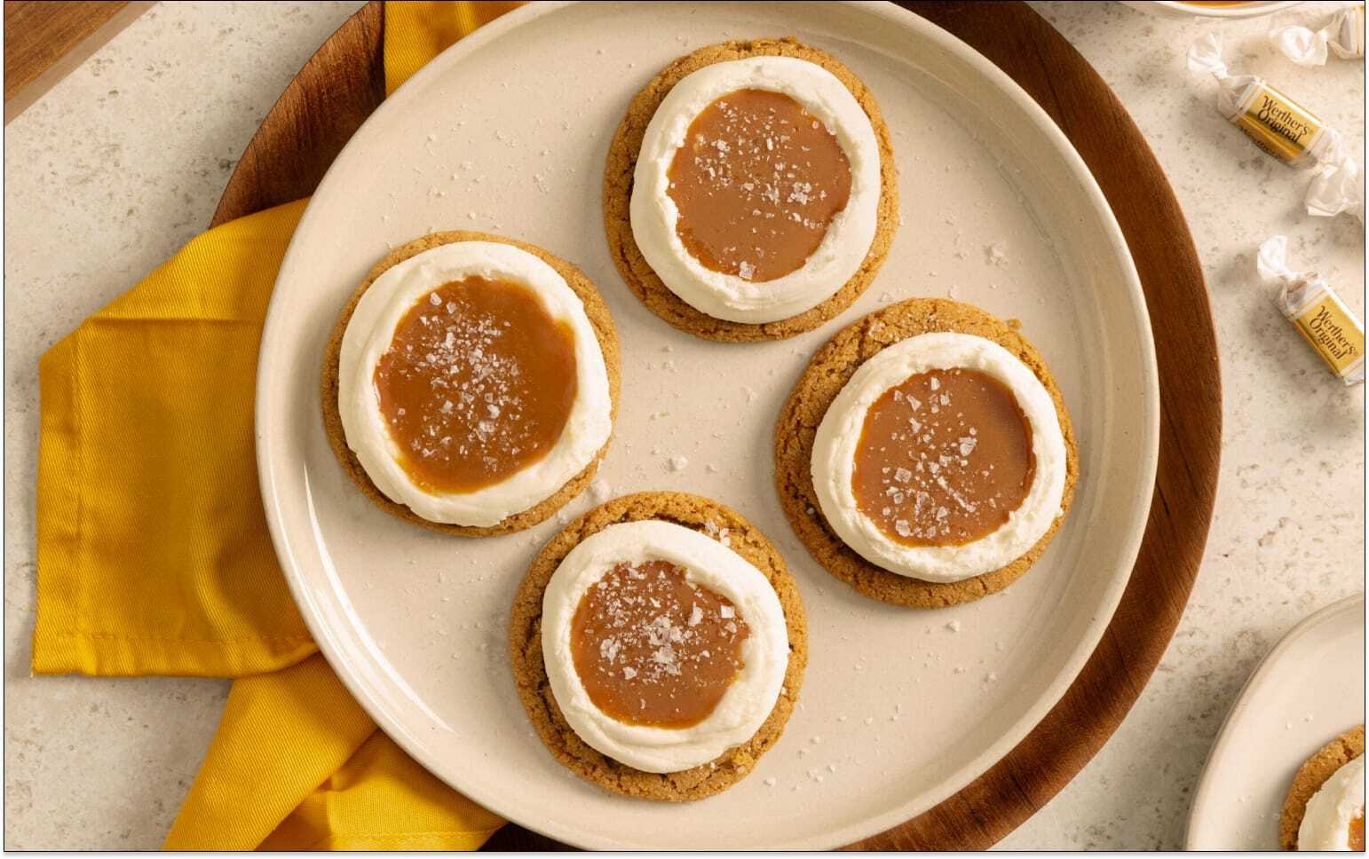 Four round biscuits topped with a ring of white icing and a caramel centre, lightly sprinkled with sugar, arranged on a cream plate with a yellow napkin underneath.